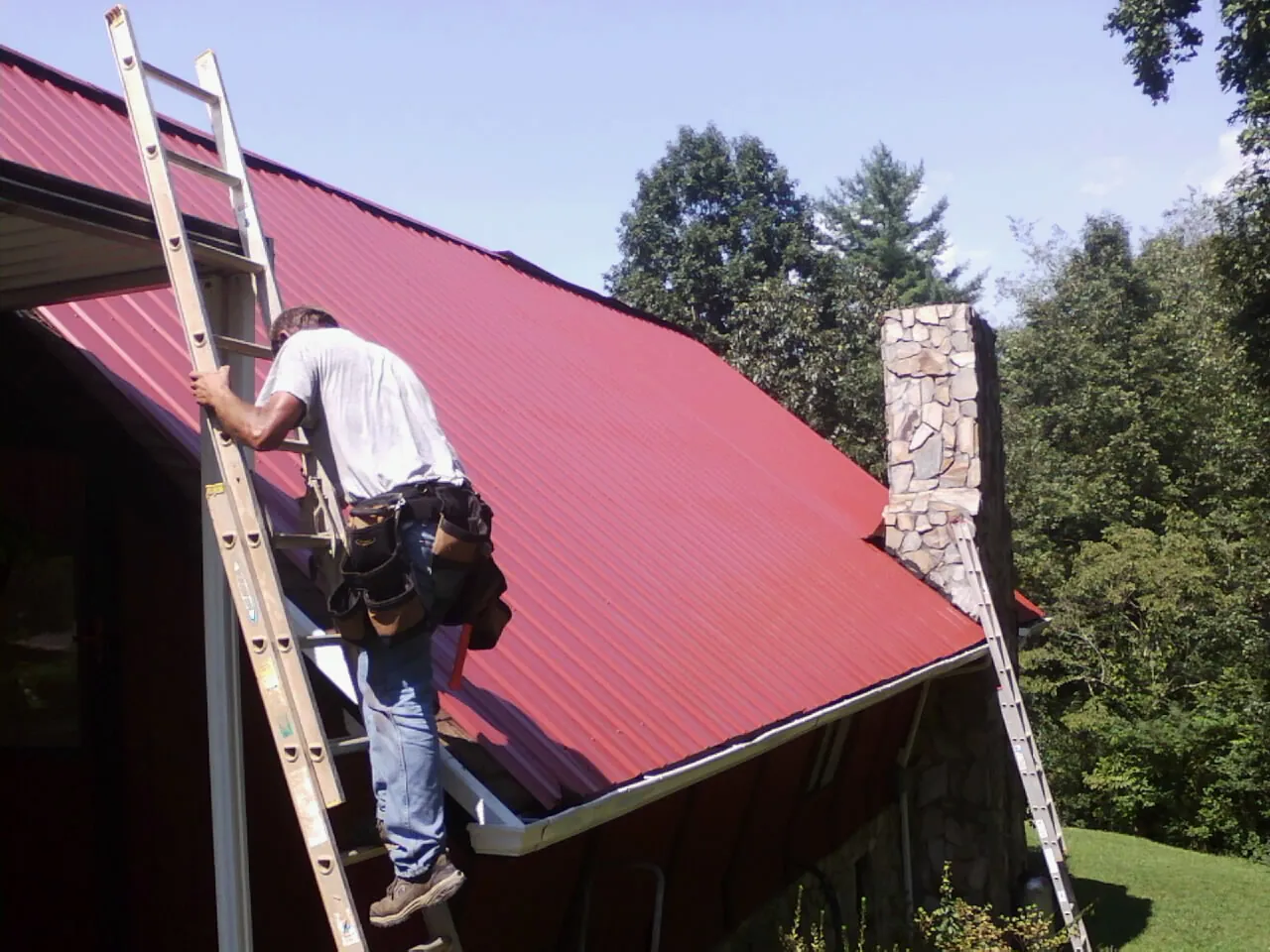Roofer on a ladder beside a red metal roof and stone chimney — Soffit & Fascia Repair by Roofing Brothers in Herndon